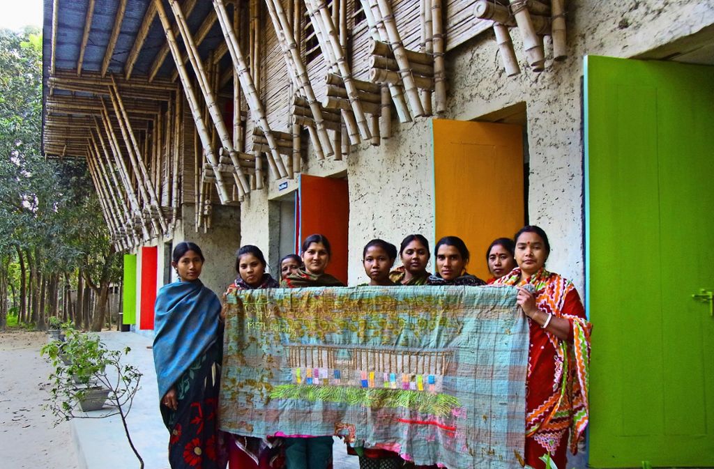 Die Frauen von Rudrapur präsentieren vor der Meti-Schule eine Decke, die mit der Ansicht der von Anna Heringer entworfenen Schule bestickt ist.