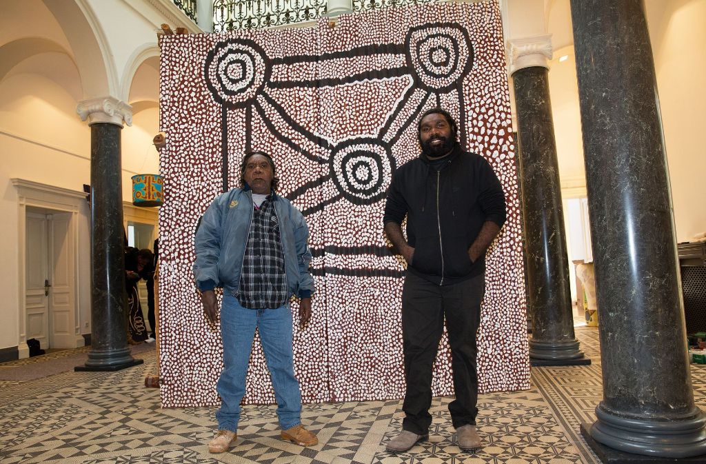 Neil Jupurrula Cooke (links) und Maxwell Tapanangka Tasman vor einem großen Bild, das verschiedene australische Traumwege symbolisiert, wie den Weg des Feuers oder des Wassers.
