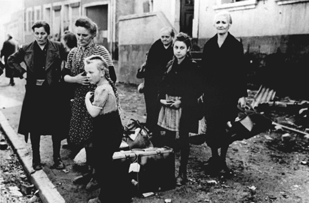 Frauen und Kinder stehen im Jahr 1945 mit ihrem Gepäck am Straßenrand und warten auf eine Transportmöglichkeit.