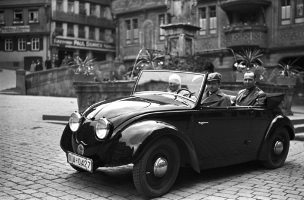 Das Werksfoto aus dem Jahr 1936 zeigt Ferry Porsche am Steuer des zweiten Volkswagen-Prototypen (V2), fotografiert auf dem Marktplatz in Tübingen. Neben ihm seine Frau Dorothea. Auf dem Rücksitz Hellmuth Zarges, ein Freund der Familie Porsche.