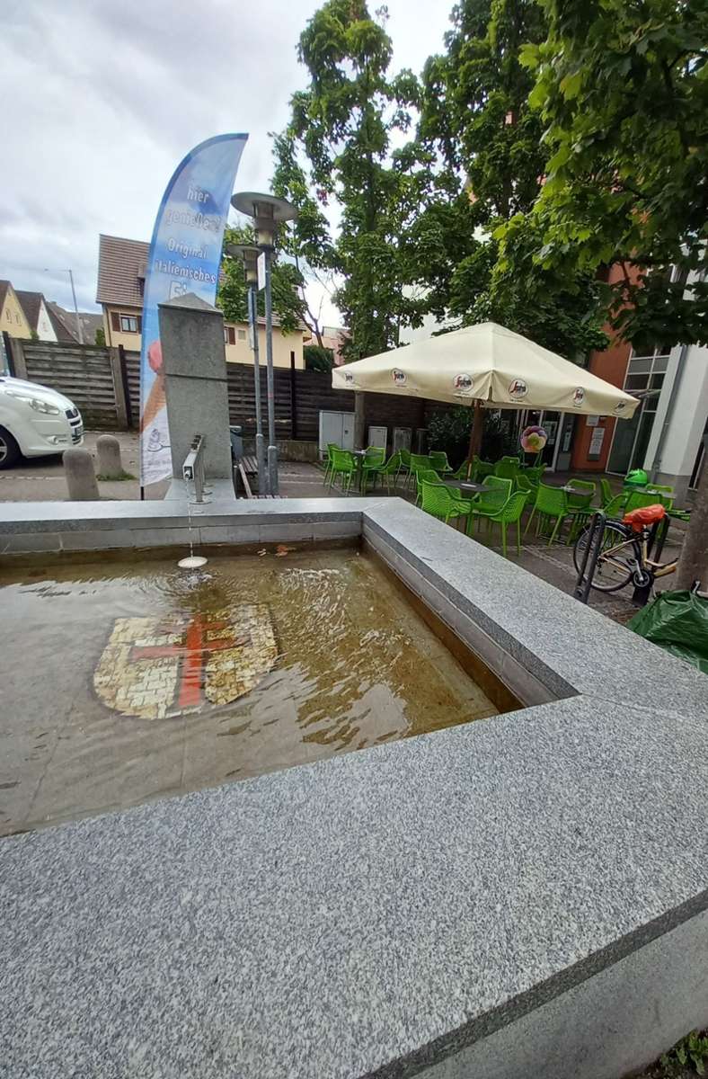 Der Brunnen am Platz ist mit dem Oeffinger Ortswappen versehen, im Hintergrund die Außenbestuhlung des neuen Eiscafés.