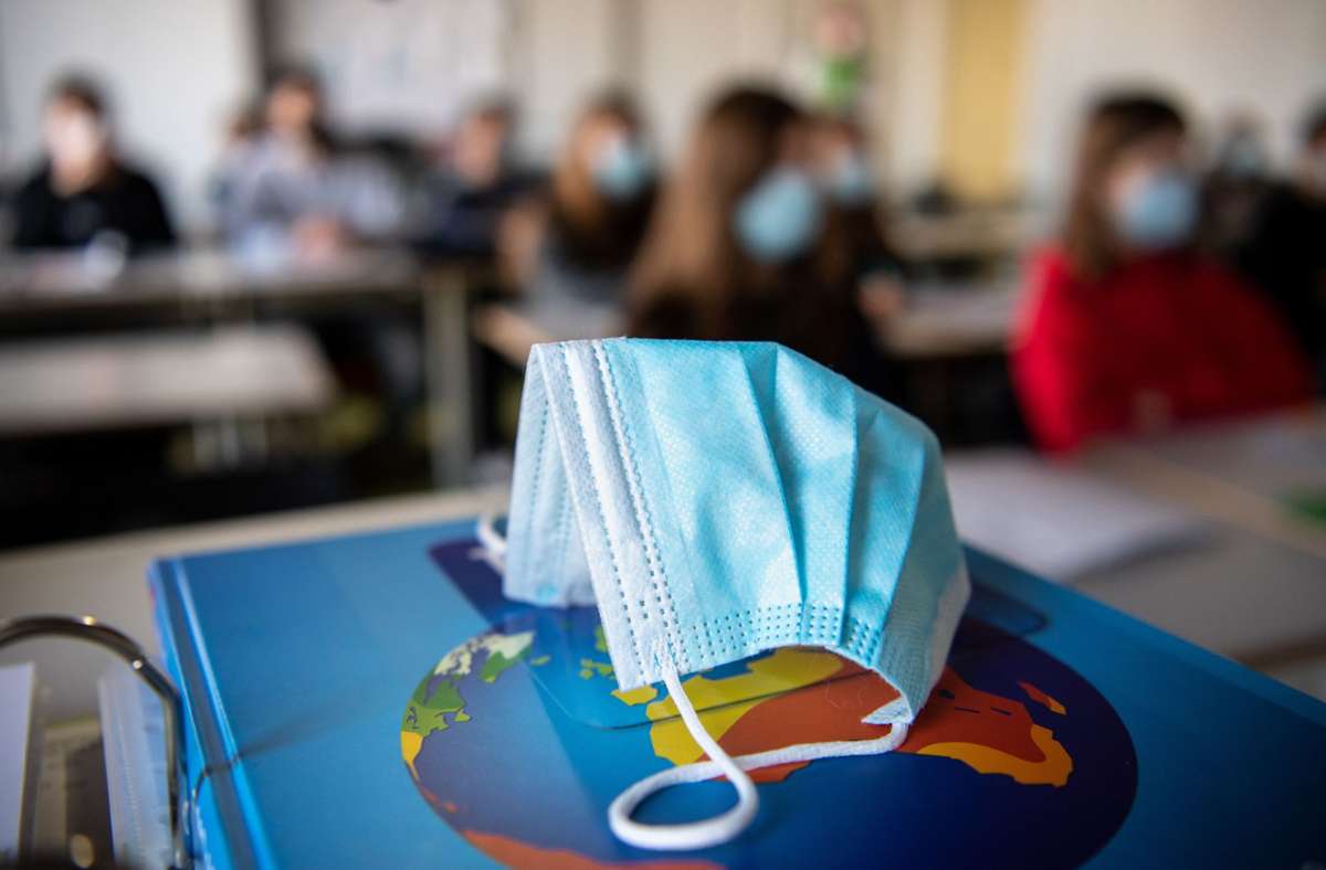 Ein Mund- und Nasenschutz liegt im Unterricht in einem Erdkunde-Unterricht, während im Hintergrund die Schülerinnen und Schüler mit Mund- und Nasenschutz zu sehen sind. Foto: Matthias Balk/dpa