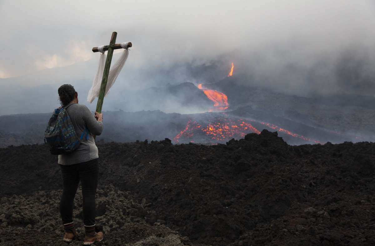 Mit einem Holzkreuz in der Hand schaut eine Frau in Richtung Vulkan.