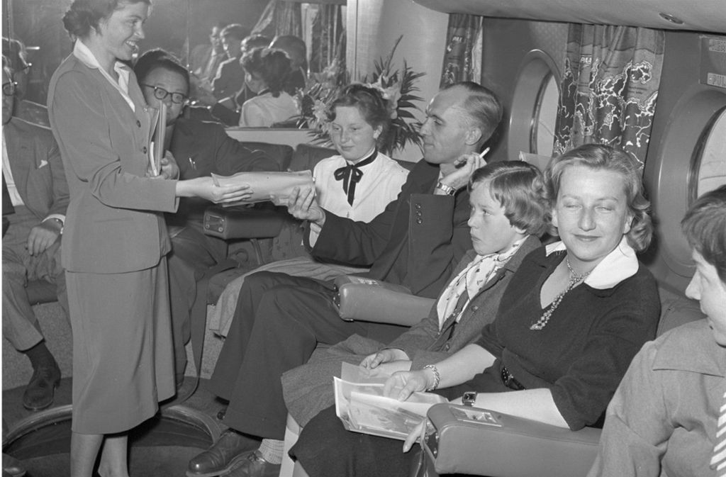 Fluggäste der 1. Klasse im Jahr 1957 auf dem Weg von München nach New York. Die Reisenden verweilen während des Fluges an einer Bar – ein Mann zieht genüsslich an seiner Zigarette, während er von einer Stewardess bedient wird.