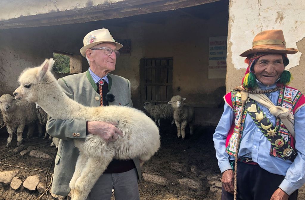 Ernst steht vor einem Stall mit Schafen und hat ein Alpaka-Cria auf dem Arm. Neben ihm in traditioneller peruanischer Kleidung der Hirte Modesto.