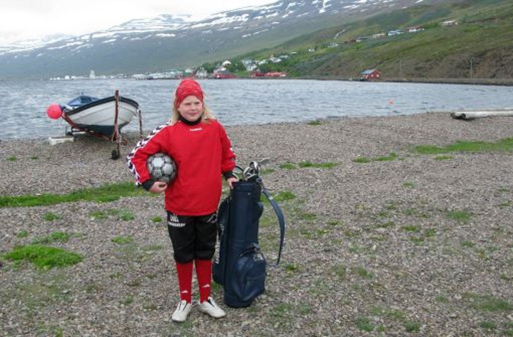 Die elfjährige Kristin aus Eskifjördur in Island steht mit Fußball und Golftasche bepackt am Fjord.