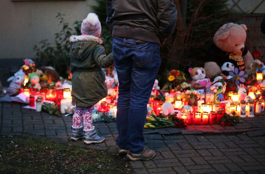 Ein Mädchen steht mit ihrem Vater vor Teddybären, Blumen und Kerzen, die in Hernevor einem Haus in einem Vorgarten liegen.