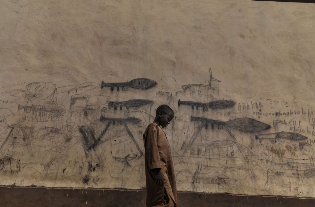 Marco Gualazzini zeigt ein Weisenkind einer Flüchtlingsgruppe am Tschadsee, das vor einer mit Waffenzeichnungen bemalten Wand läuft.
