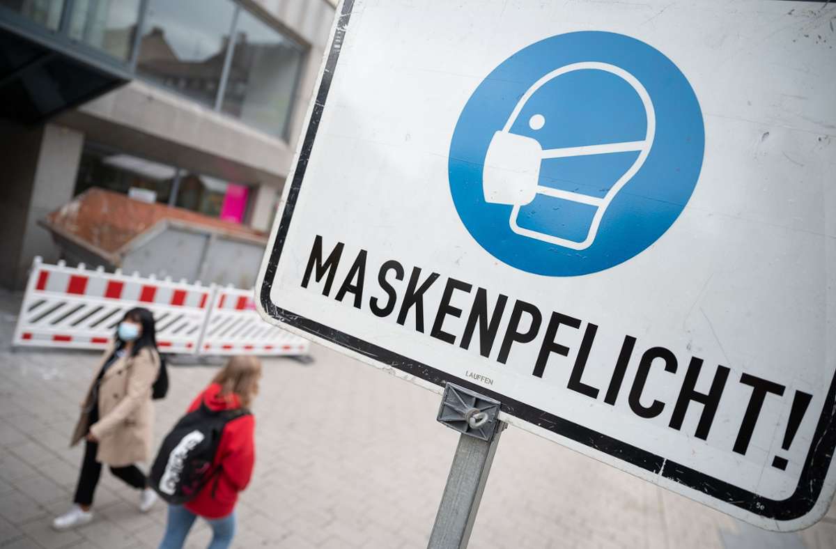 Zwei Passantinnen gehen in einer Fußgängerzone hinter einem Schild vorbei, auf dem „Maskenpflicht“ steht.