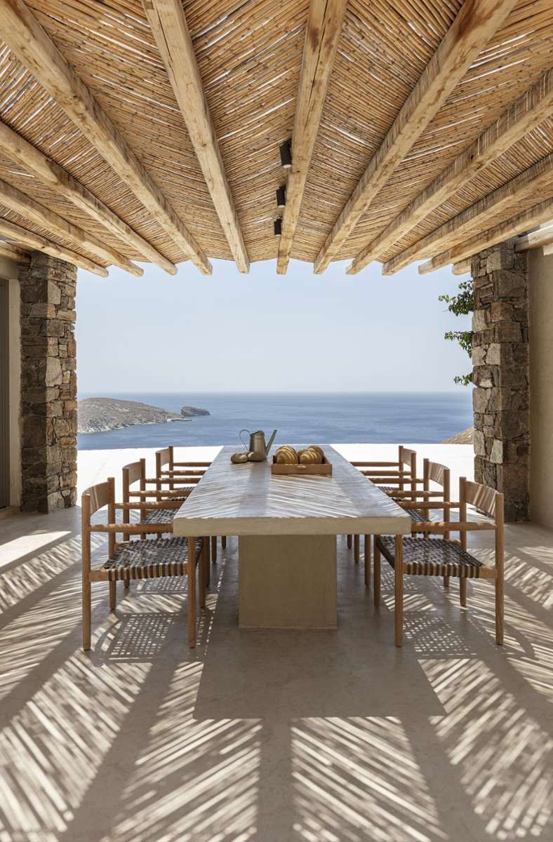 Sommerhaus Xerolithi von Sinas Architects in Serifos, Griechenland. Die Bambus-Pergola schützt vor der Sonne und sorgt für schöne Muster auf dem Tisch und dem Boden, so lässt sich die Aussicht aufs Meer schattig genießen. Da auch im Innenraum Bambus an der Decke zu finden ist, gestaltet sich der Übergang vom Innenraum zum Außenraum harmonisch.
