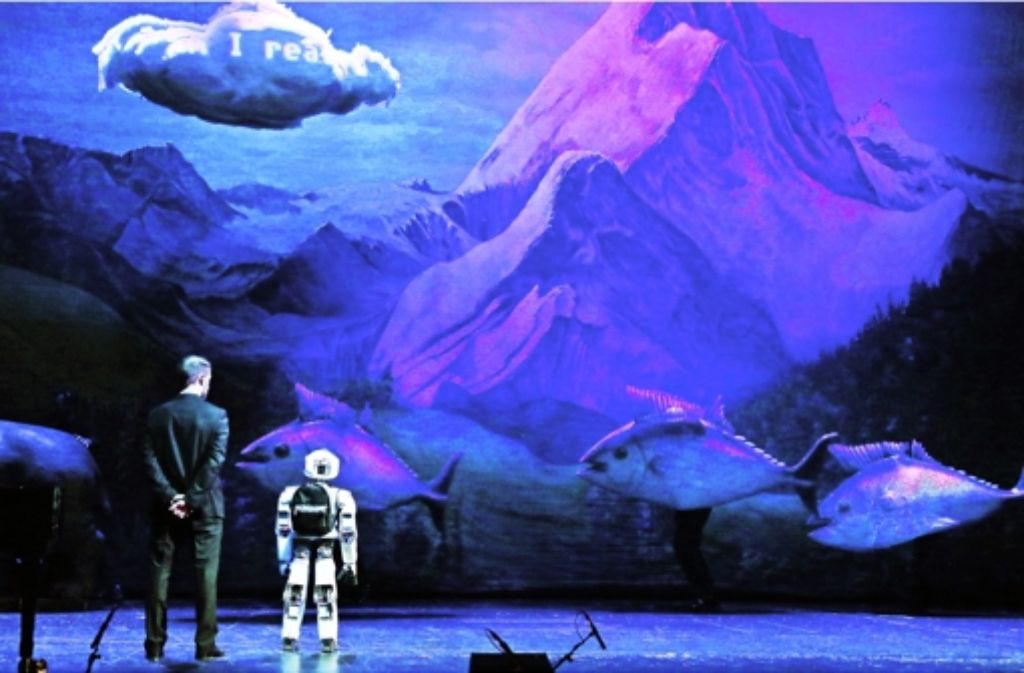Ob ihm gefällt, was er sieht? Der Roboter Myon beobachtet das Bühnenbild in Berlin: Fische vor Bergkulisse. Am I real?, fragt die Projektion auf der Wolke. In einer Bildergalerie zeigen wir weitere Eindrücke von der Oper „My Square Lady“.