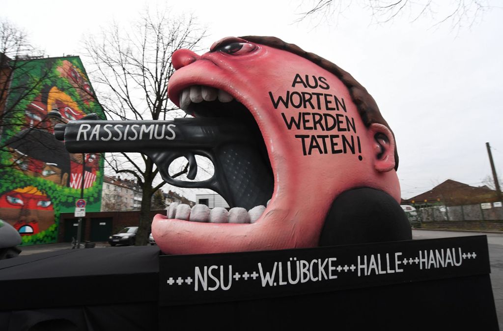 Düsseldorf: Eine Pistole mit der Aufschrift „Rassismus“, die aus dem Mund eines Mannes mit hochrotem Kopf ragt, mahnt nach dem Anschlag von Hanau. Auf seiner Wange steht: „Aus Worten werden Taten!“
