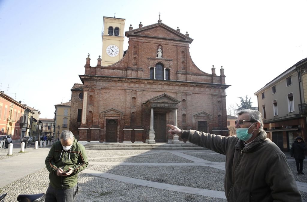 Zwei Männer Menschen mit Mundschutz stehen vor der Kirche San Biagio. In Italien soll die weitere Ausbreitung des Coronavirus mit drastischen Mitteln bekämpft werden.