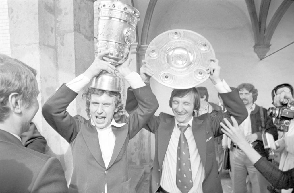 Saison 1977/78: Das Preisschießen. Kann Gladbach 11 Tore schießen? Es kann. Es hört gar nicht mehr auf. „Am Ende hat der Schiedsrichter die Bälle aus dem Tor geholt. Wir konnten nicht mehr, wir hatten da Bleigewichte an den Füßen,“ erinnert sich BVB-Libero Lothar Huber. Doch sieben Tore aufzuholen, reicht den Gladbachern nicht: Zehn hätten es sein müssen, mit 12:0 besiegen sie die Borussia. Köln rettet sich durch ein 5:0 im Hamburger Volksparkstadion gegen den FC St. Pauli, der nicht am Millerntor spielte, über die Ziellinie. DFB-Ermittlungen gegen die Dortmunder verlaufen im Sande, der damalige BVB-Coach Otto Rehhagel wird zur „Otto Torhagel“ und von den Schwarz-Gelben einen Tag nach der Pleite in Düsseldorf entlassen. Die Kölner werden derweil begeistert von ihren Fans in der Innenstadt empfangen. Die Spieler Heinz Flohe (im Foto links) und Hannes Löhr präsentieren Kölns Trophäen – den Pokal und die Meisterschale.