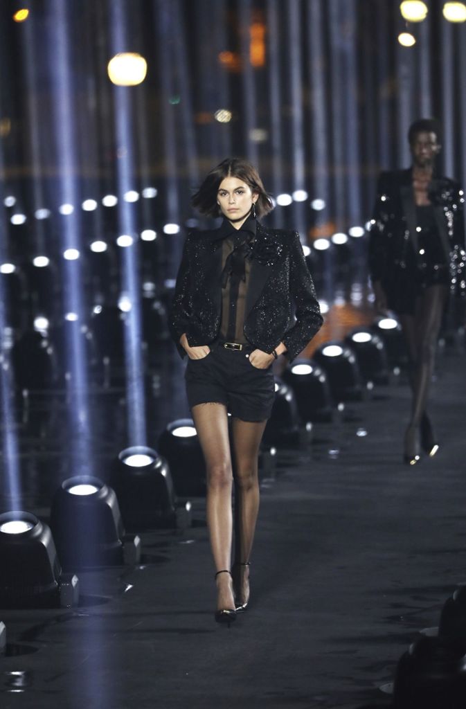 Und auch sie lief bereits auf anderen Fashion-Weeks diese Saison und ist auch darüber hinaus bekannt: Kaia Gerber, die Tochter des ehemaligen Models Cindy Crawford, in schwarzer durchscheinender Schleifchen-Bluse mit schwarzer Hotpant und Glitzer-Blazer auf dem Catwalk der Saint-Laurent-Show.