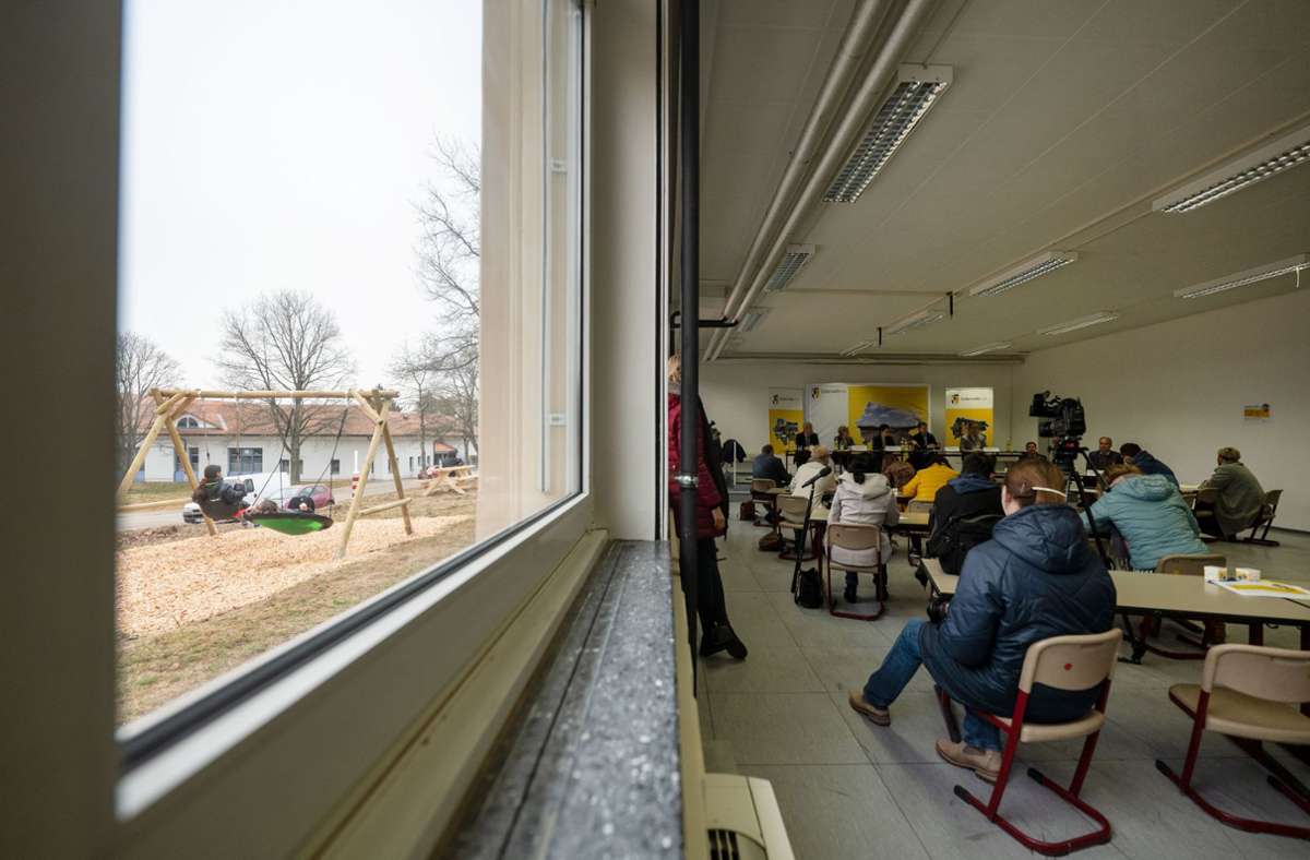 Journalisten nehmen in der ehemaligen Zollernalb-Kaserne an einer Pressekonferenz in einem improvisierten Schulsaal teil, während draußen geflüchtete Kinder spielen.